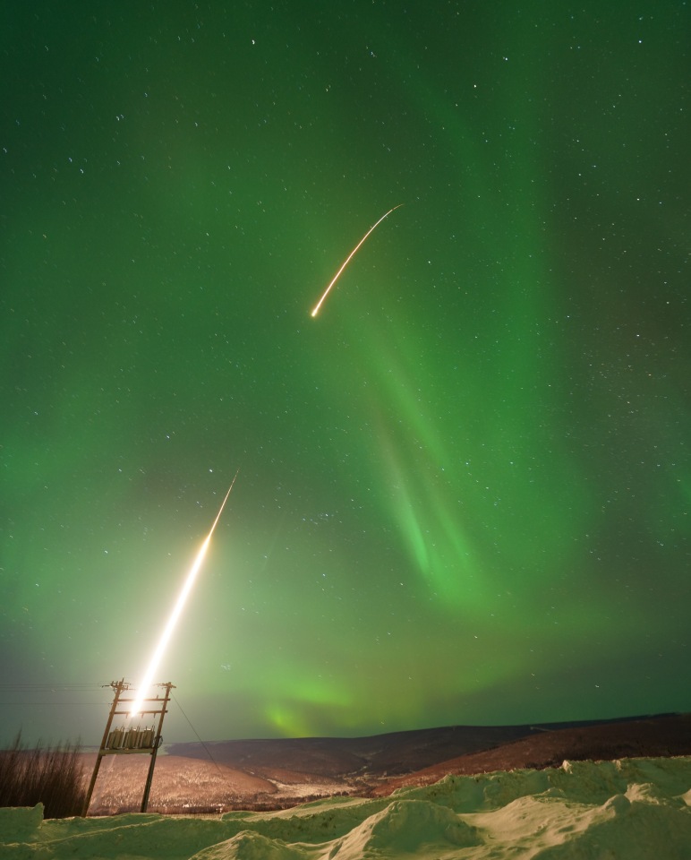A long-exposure nighttime photograph shows a sounding rocket launching upward, leaving a bright curved streak of light against a green aurora-filled sky. Stars are visible overhead, with snow-covered ground and utility poles in the foreground.