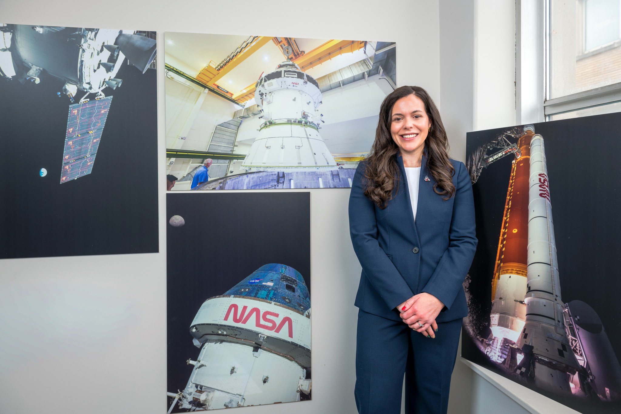 A professionally dressed woman stands in a gallery style room surrounded by large NASA photographs showing the Orion spacecraft in space, engineers working on a module, a close-up of the Orion spacecraft with the Moon behind it, and the SLS (Space Launch System) rocket illuminated on the launch pad.