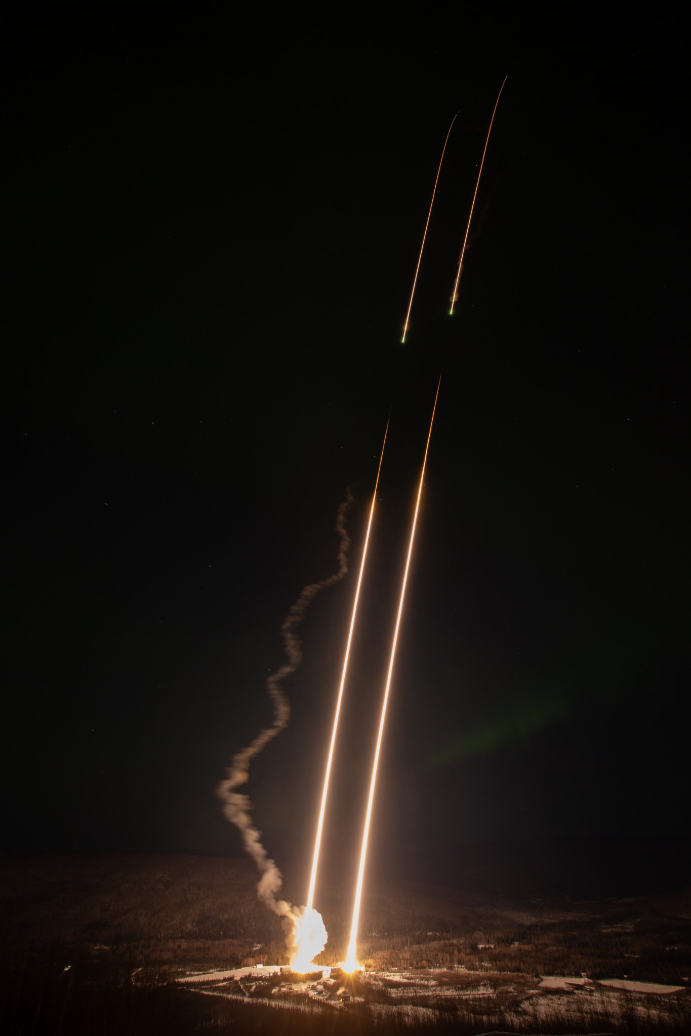 A long-exposure nighttime photograph shows two sounding rockets launching side by side, leaving parallel bright streaks of light rising into a dark sky. A faint smoke plume trails behind one rocket, with the launch pad and surrounding snowy landscape visible below.