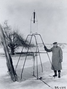On a snowy March 16, 1926, Dr. Robert H. Goddard rests his hand on the testing frame supporting his liquid fuel rocket at Ward Farm in Auburn, Massachusetts. A wooden door is propped up at an angle next to the frame where Goddard’s assistant, Henry Sachs, later sheltered after lighting the rocket.