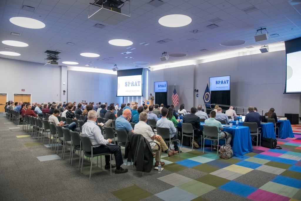 People seated in a large auditorium listening to a presentation.