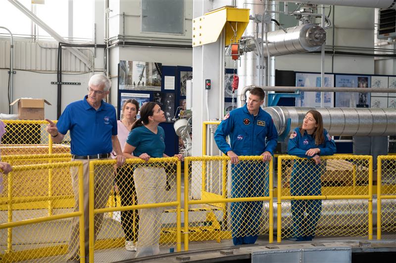 A small group stands inside NASA’s Neil Armstrong Test Facility, with two people in blue NASA flight suits and others in professional clothing, surrounded by industrial equipment, pipes, and yellow safety railings. NASA/Sara Lowthian-Hanna