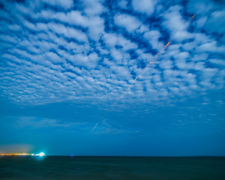 An orange and white streak cuts diagonally through a cloudy sky and down to Earth in the distance. The clouds have an unusual, rippling, wave-like pattern.
