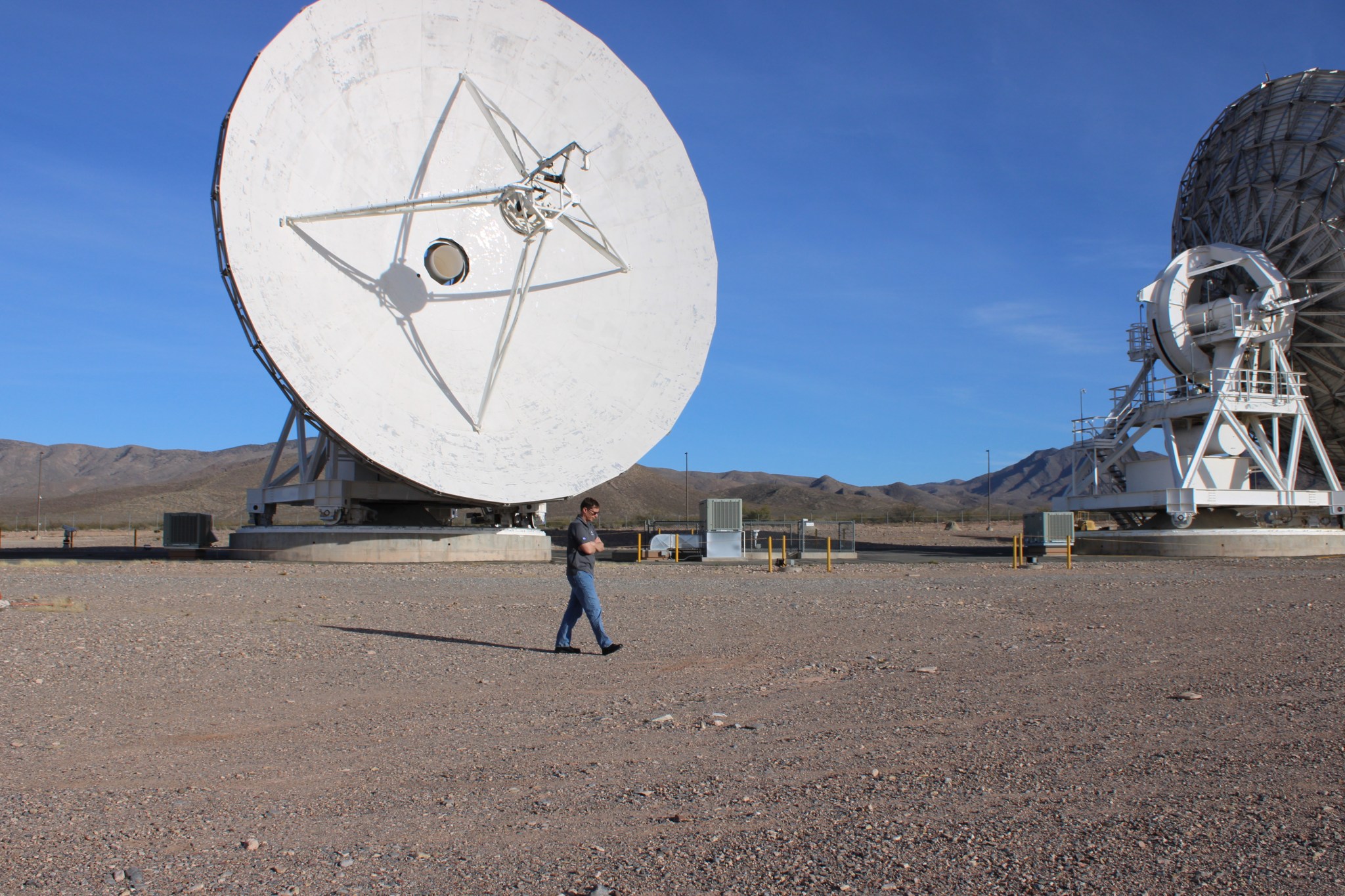 A man walks across a gravel lot between two large satellite dish antennas in a desert landscape with mountains and a clear blue sky.