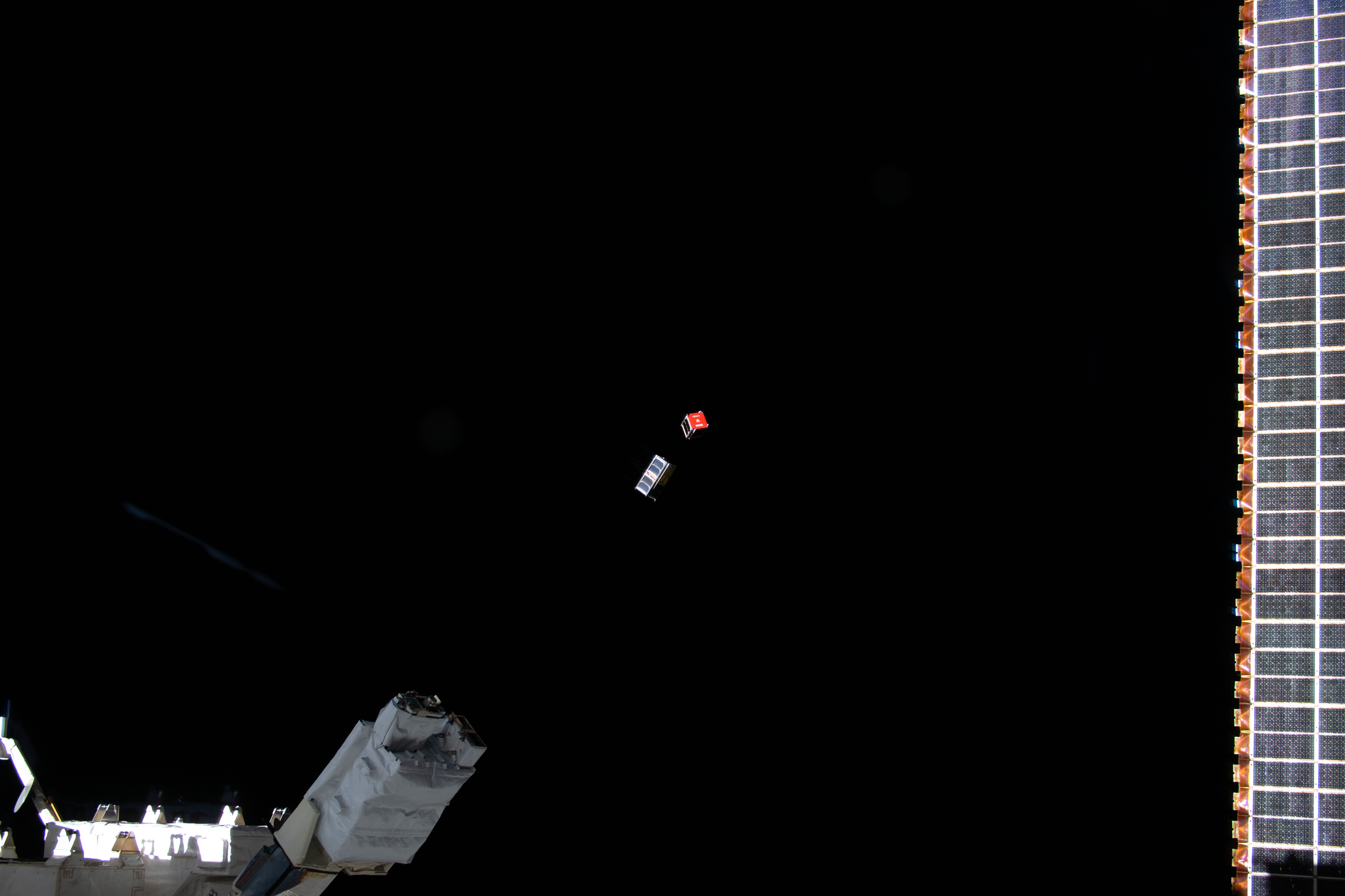 Two CubeSats, one silver and one with a red face, are ejected from the International Space Station (bottom left) into the blackness of space.
