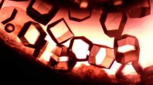 An extreme close-up on crystals. They are geometric in shape, some looking like squares while others are hexagons. Orange light shines from behind the crystals. The light shines through the crystals that face us and highlights their edges.