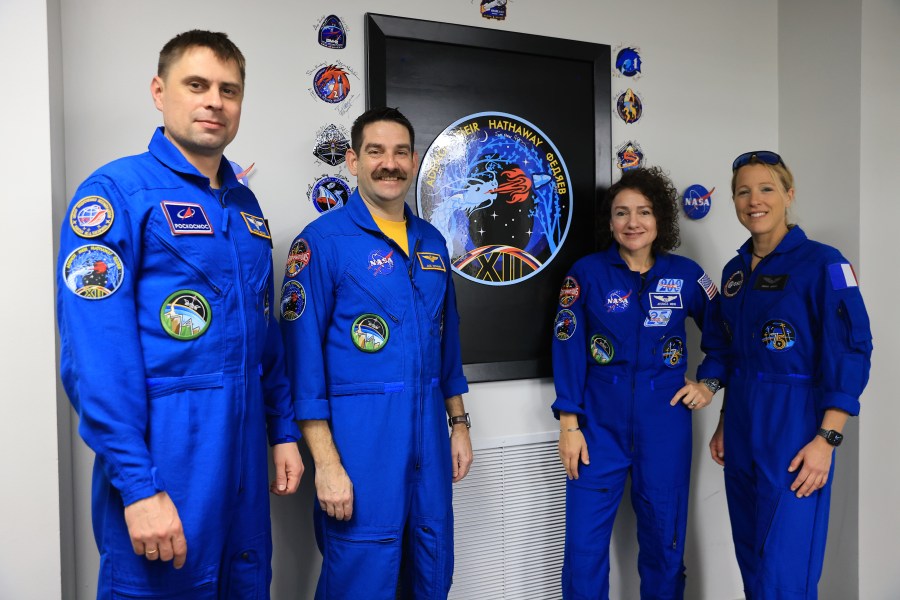 Four people - two men on the left and two women on the right - pose with the Crew-12 mission insignia. They are all wearing blue jumpsuits with various patches on them. The insignia is on the wall, framed in a black recess. Autographed patches are stuck on the wall around the black frame.