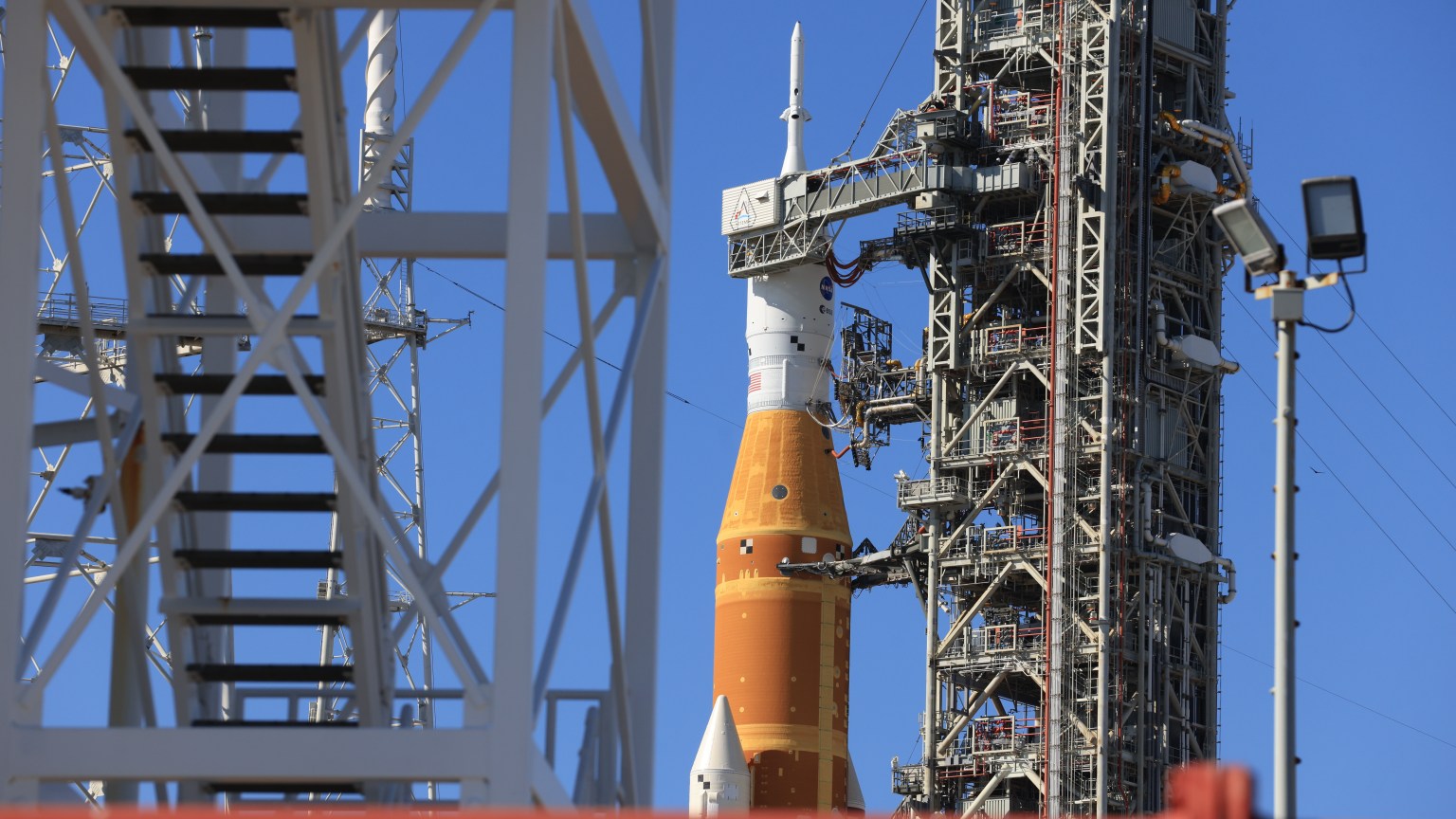 A side view of the launch pad where the Artemis II SLS rocket and Orion spacecraft sit on top of the mobile launcher. It shows one of the white twin SLS solid rocket boosters, orange core stage, and Orion spacecraft.