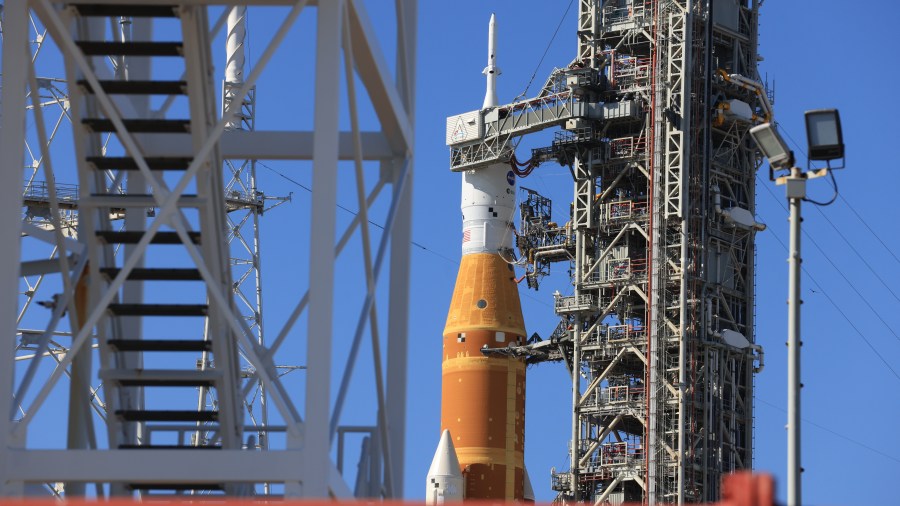 A side view of the launch pad where the Artemis II SLS rocket and Orion spacecraft sit on top of the mobile launcher. It shows one of the white twin SLS solid rocket boosters, orange core stage, and Orion spacecraft.