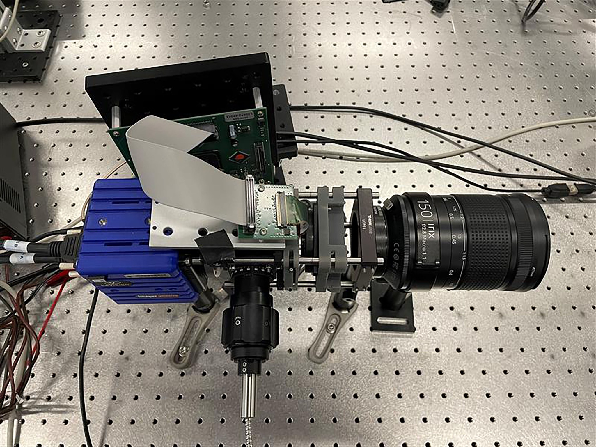 A black camera lenses is mounted along with a black, sliver, and blue set up to stabilize the camera. The system is standing on a silver table with even distributed holes.