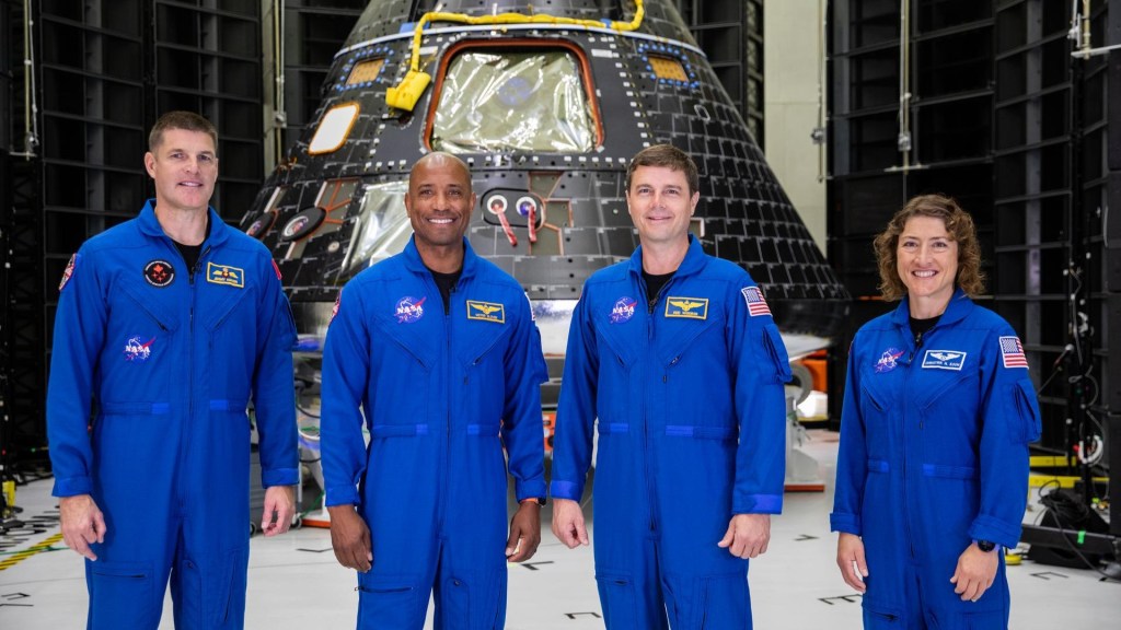 A picture of four astronauts in blue flight suits standing in front of the Orion capsule.