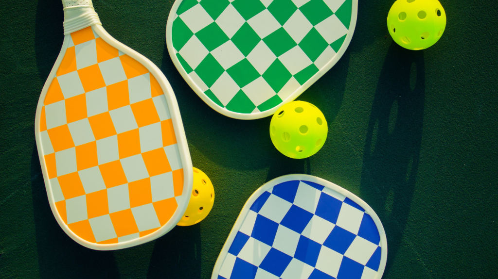 Three pickleball paddles with checkered patterns in orange, green, and blue, alongside three yellow pickleballs on a dark green surface.