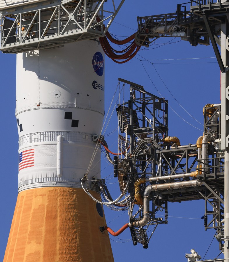 A section of a rocket seen with multiple lines, or umbilicals, connecting it to eqiupment on a tower.
