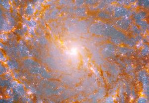 A close-up view shows a galaxy’s bright, yellow center, surrounded by patches of glowing orange-brown dust and pink stars.