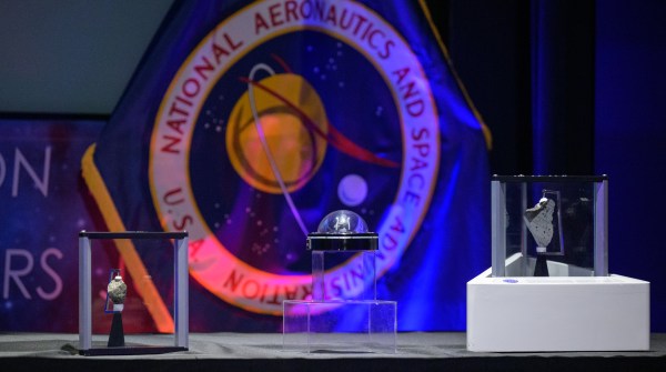 Three Moon rocks are encased in three different kinds of containers. They all sit on a flat surface. Behind them is the NASA flag with the agency's seal on it. The flag is lit from the bottom by red and blue lighting.