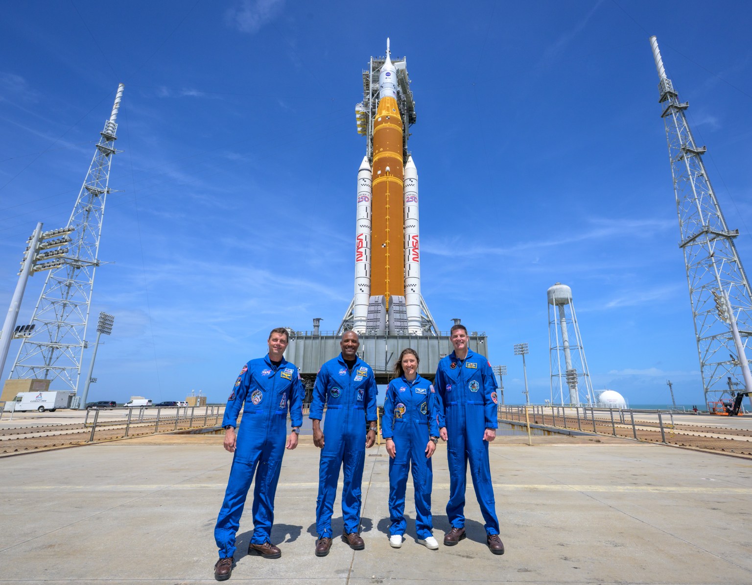 Four NASA Artemis II astronauts at the launch pad with the SLS rocket in the back.