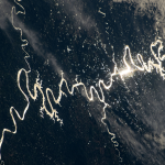 Sunlight can be seen glinting off the winding rivers across the Pampas grasslands of South America. The light forms a long winding shape from right to left across a dark blue background with patches of white clouds.