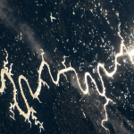 Sunlight can be seen glinting off the winding rivers across the Pampas grasslands of South America. The light forms a long winding shape from right to left across a dark blue background with patches of white clouds.
