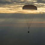 A picture of a capsule attached to a parachute during the return of a mission with the sky in the background.