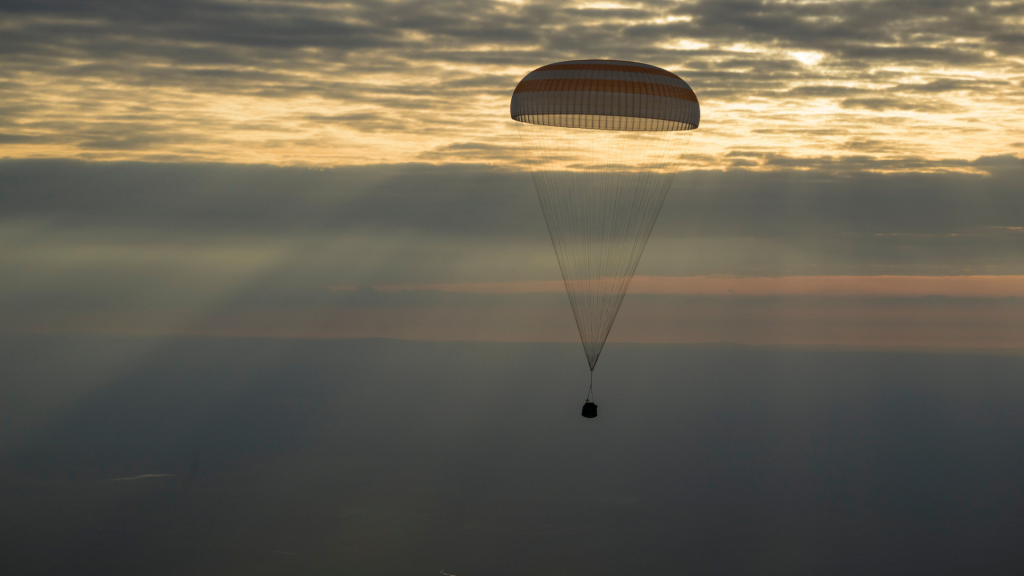 A picture of a capsule attached to a parachute during the return of a mission with the sky in the background.