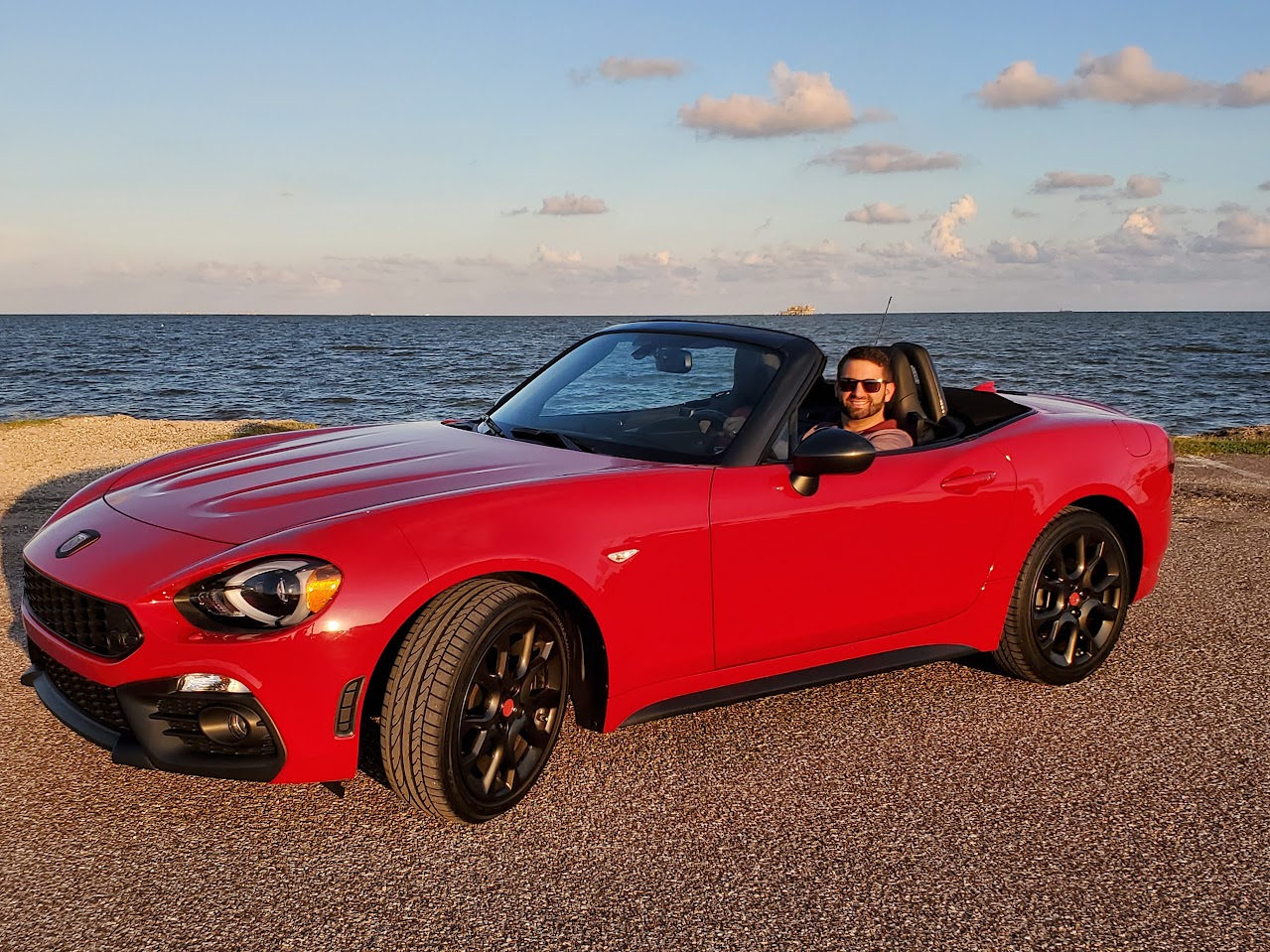 A man poses inside of a red convertible by the beach.