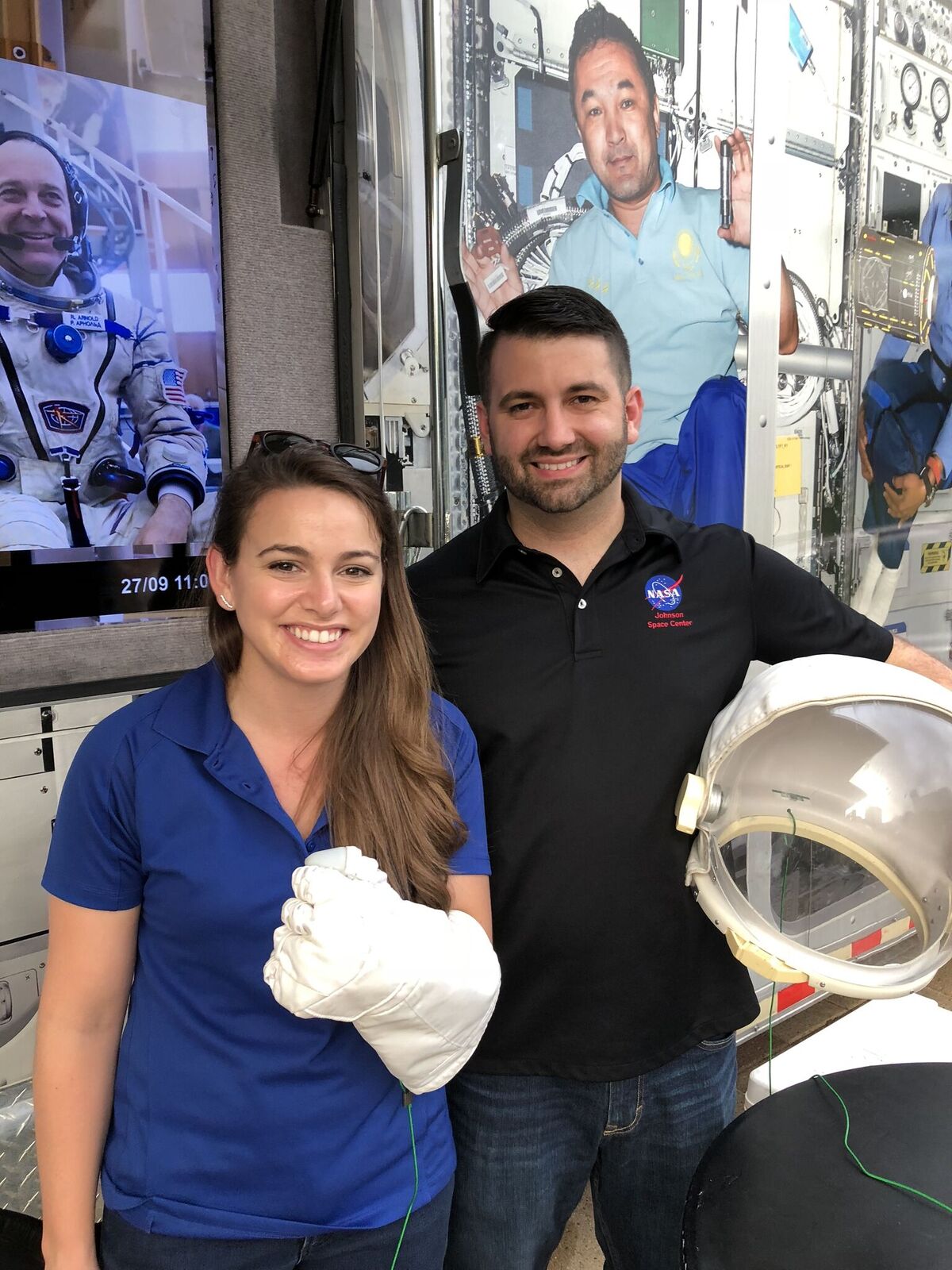 Two people pose in front of a portrait of an astronaut. The person on the left is giving a thumbs up wearing a spacesuit glove. The person on the right