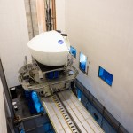 A large white NASA flight simulator dome, bearing the NASA logo, mounted on a motion platform inside a tall industrial facility with a linear track system below.