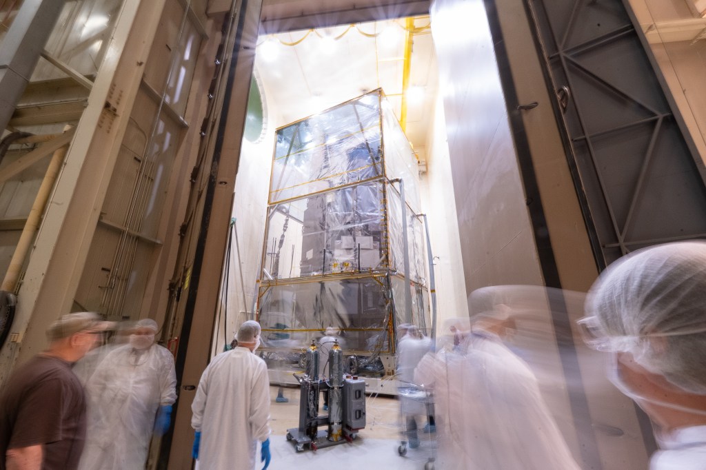A creative style photo capturing movement of people in white gowns. Their appearances are blurred and look almost spectral. Behind them through an open doorway, the Roman observatory sits in a tall room. The observatory is encased in a clear protective structure.