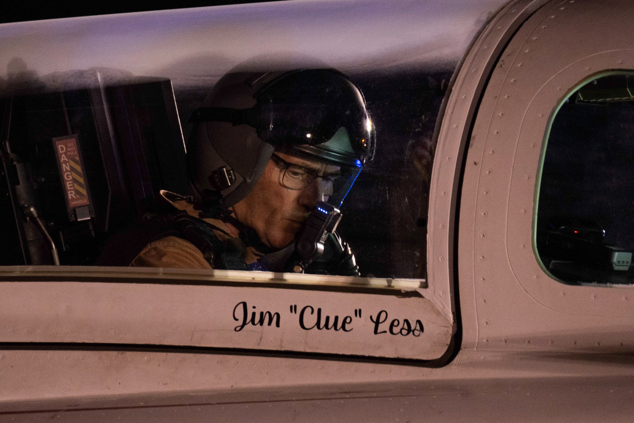 NASA test pilot Jim “Clue” Less sits in the X-59's cockpit in a close-up photo. Less is wearing his flight suit, a flight helmet, and glasses, with a radio pressed close to his face. His name and call sign are written just under the canopy glass.