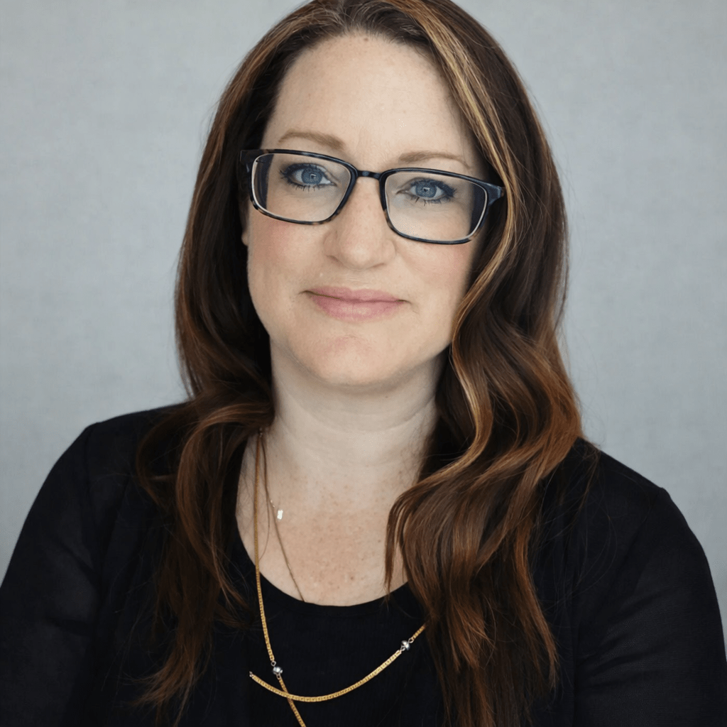 A woman poses for a portrait image. She has dark hair, blue eyes and is wearing glasses.