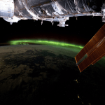 A vibrant green aurora is visible on the arched horizon of the Earth. A portion of the space station is visible on the top part of the picture and one of the space station’s solar array wings is in front of the Earth.