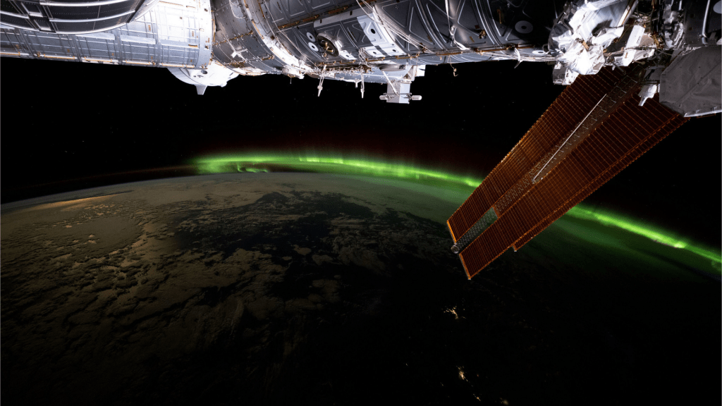 A vibrant green aurora is visible on the arched horizon of the Earth. A portion of the space station is visible on the top part of the picture and one of the space station’s solar array wings is in front of the Earth.