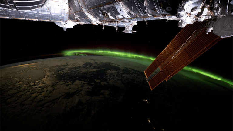 A vibrant green aurora is visible on the arched horizon of the Earth. A portion of the space station is visible on the top part of the picture and one of the space station’s solar array wings is in front of the Earth.