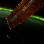 A vibrant green aurora is visible on the arched horizon of the Earth. A portion of the space station is visible on the top part of the picture and one of the space station’s solar array wings is in front of the Earth.