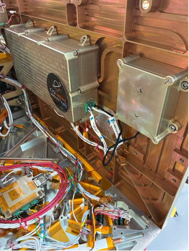 Two silver metal boxes are mounted to a copper-colored panel. The box on the left has a CisLunar Industries logo on it. Below the mounted boxes are multi-colored wires.