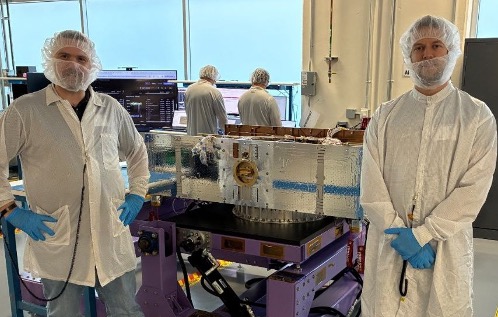 Two researchers wearing white lab coats, beard and hair nets, and blue latex gloves stand in front of a purple metal table. A flat, reflective silver panel stands on the table with a round brass-colored port in its center.