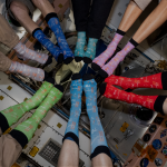 A group of people wearing different colorful socks posed in a circle with their feet all facing the center. They are floating in the picture because they are on the International Space Station on orbit.