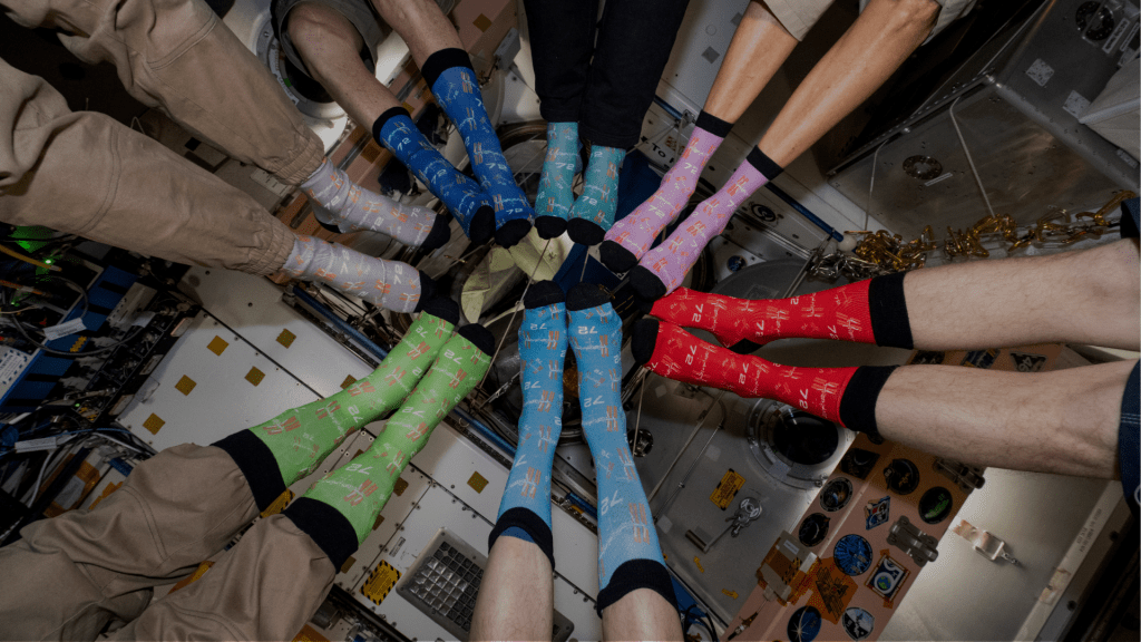 A group of people wearing different colorful socks posed in a circle with their feet all facing the center. They are floating in the picture because they are on the International Space Station on orbit.