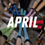 A group of people wearing different colorful socks posed in a circle with their feet all facing the center. They are floating in the picture because they are on the International Space Station on orbit.