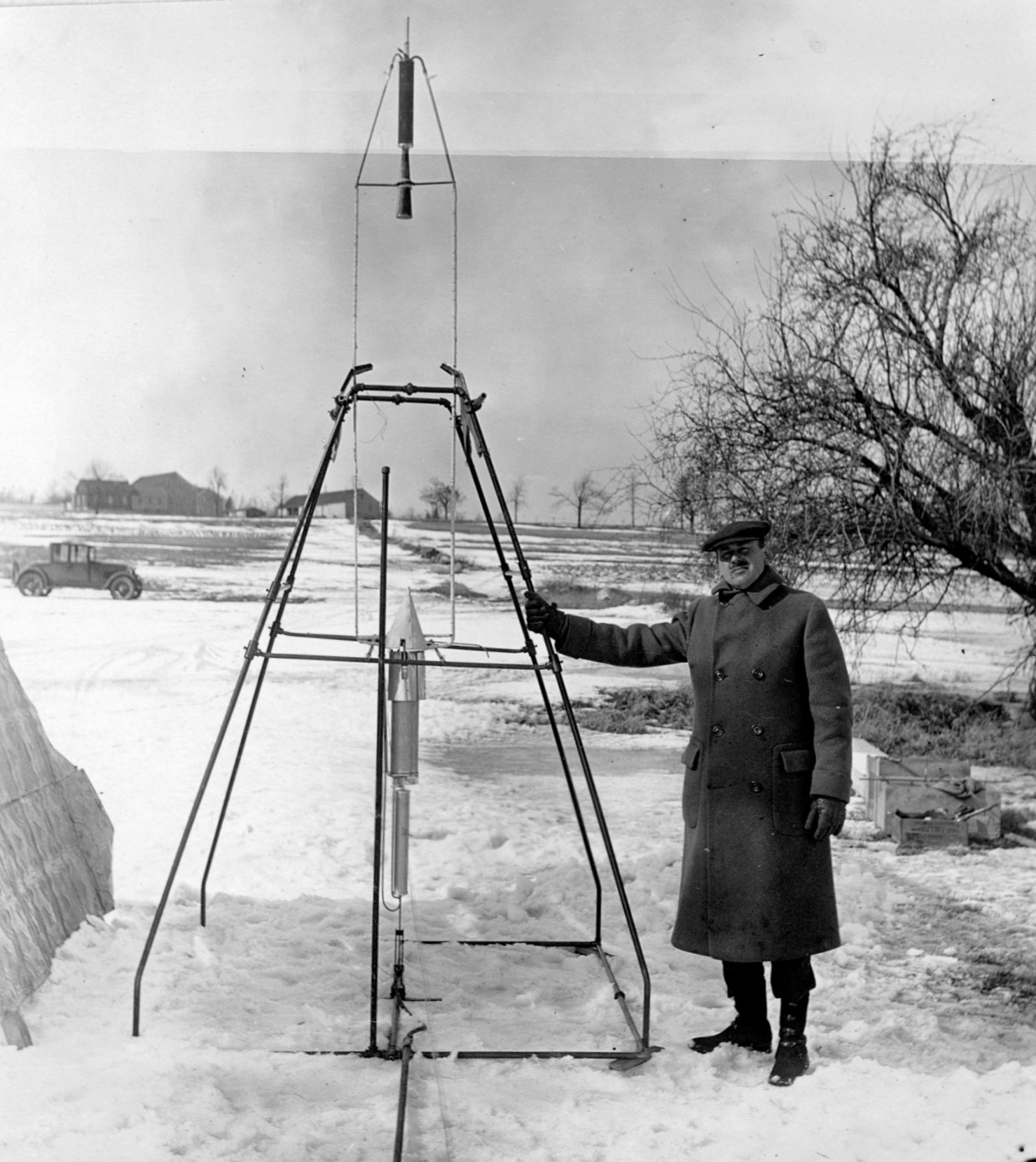 black-and-white photograph of a man in an overcoat and hat standing next to a slender, 11-foot-tall rocket supported by a metal frame