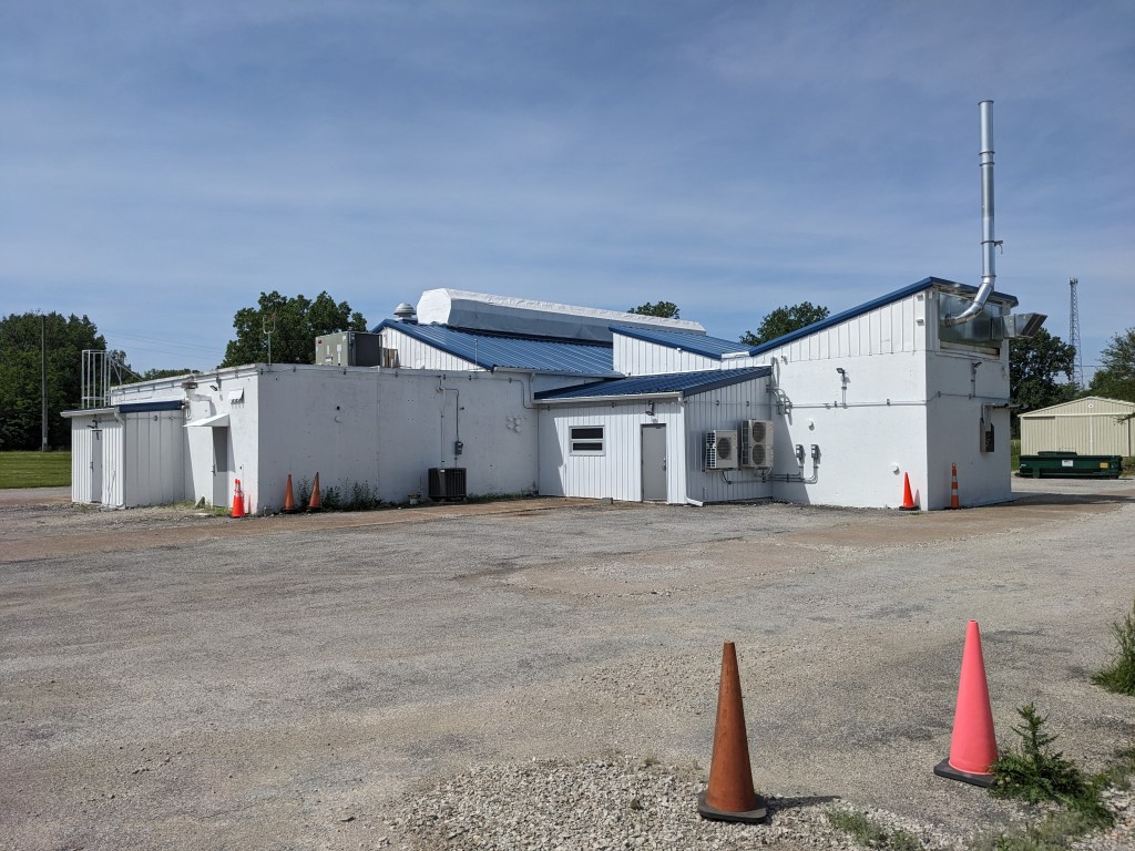 The back of a white building with a blue roof and a large metal exhaust sticking up out of the side. The surrounding parking lot is empty. Orange cones are placed near the building and in the foreground.