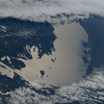 Lake Michigan beams in the afternoon sunlight as clouds blanket its southern tip—spanning Michigan, Indiana, and Illinois—in this photograph taken from the International Space Station while orbiting 263 miles above Earth. The northern tip of Lake Huron is visible at left.