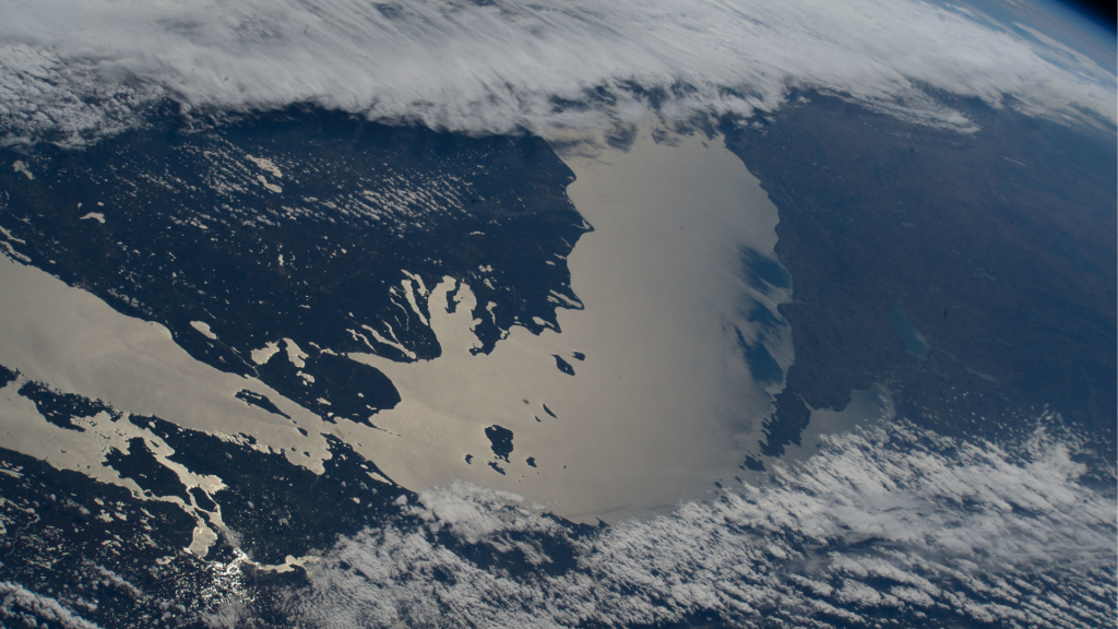 Lake Michigan beams in the afternoon sunlight as clouds blanket its southern tip—spanning Michigan, Indiana, and Illinois—in this photograph taken from the International Space Station while orbiting 263 miles above Earth. The northern tip of Lake Huron is visible at left.