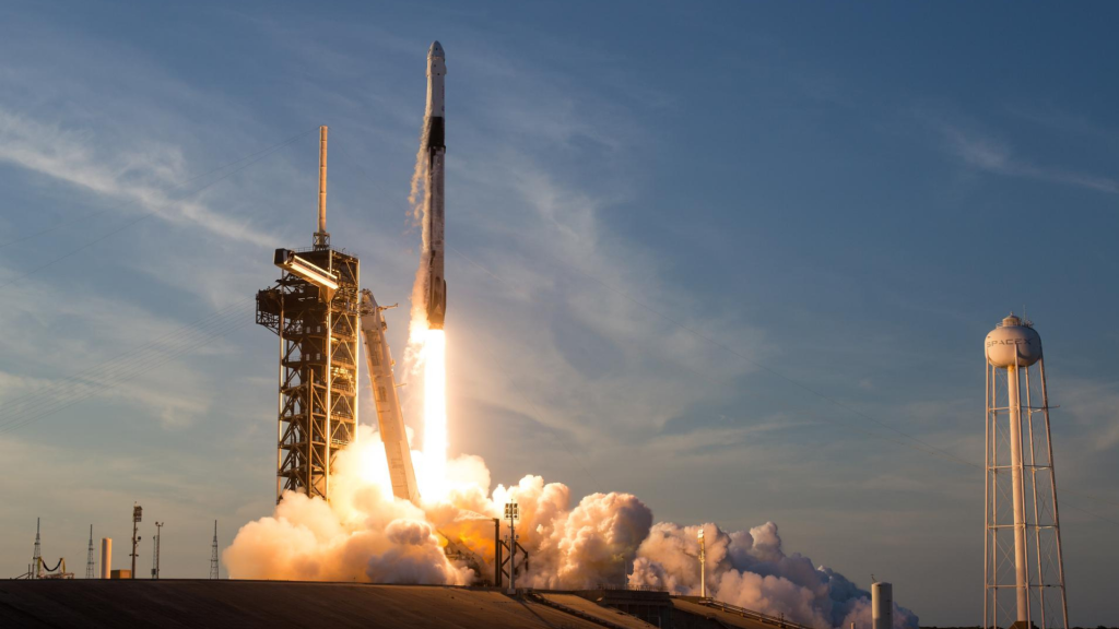 A picture of a rocket and capsule launching off the pad with plumes coming out the bottom during the daytime.