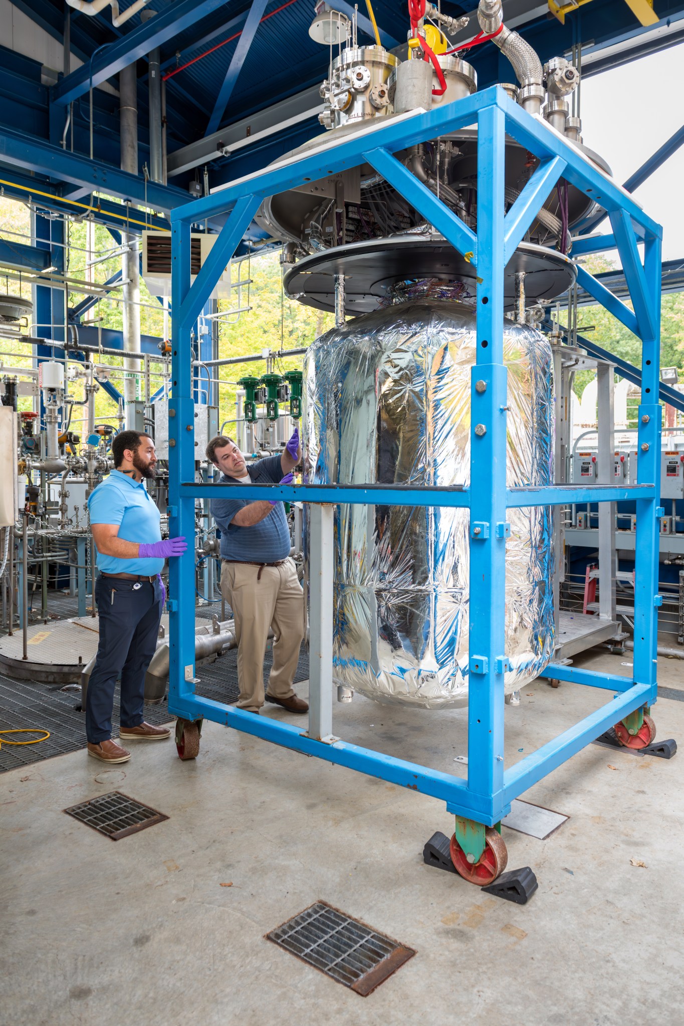 Two engineers stand beside a large stainless steel cylindrical chamber wrapped in reflective insulation, mounted inside a bright blue steel frame on wheels. One engineer points at the equipment while the other looks on. Pipe, valves, and metal structures are visible in the background.