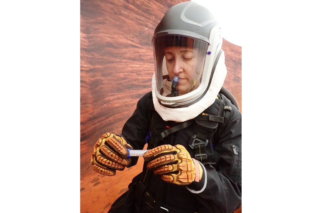 Caption: CHAPEA mission 2 medical officer, Ellen Ellis, collects samples during an extravehicular activity, also known as a spacewalk. C