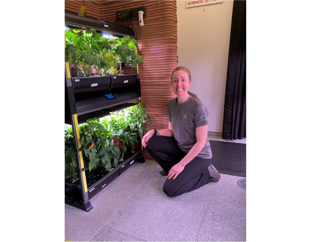 Crew Health and Performance Exploration Analog (CHAPEA) Mission 2 medical officer, Ellen Ellis, kneels in front of the hydroponic growth system.