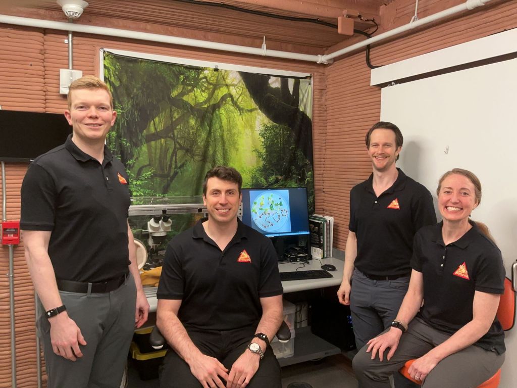 The Crew Health and Performance Exploration Analog (CHAPEA) Mission 2 crew stands in front of a science glove box, marking 300 days inside the habitat (from left: James Spicer, Ross Elder, Matthew Montgomery, Ellen Ellis).