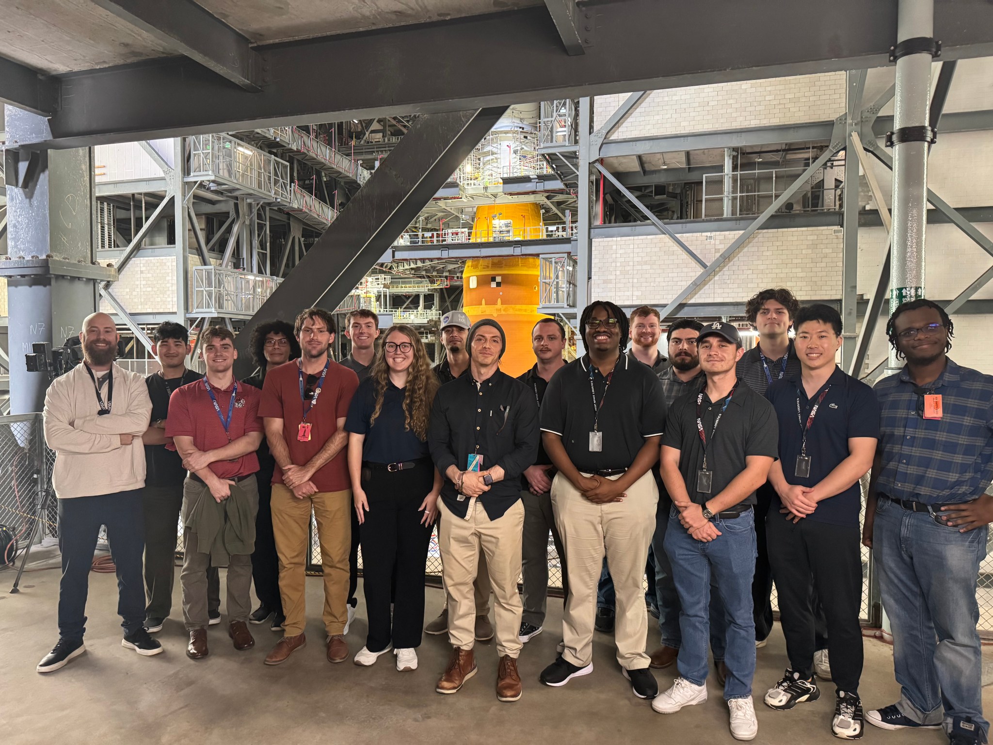 A group of approximately 18 young professionals, identified as NASA Pathways interns, posing for a group photo inside a large industrial assembly facility. They are standing on a concrete platform under a steel support structure. In the background, a massive orange rocket stage is visible within a complex gray gantry and scaffolding system. The interns are dressed in professional-casual attire and most are wearing NASA-branded lanyards.