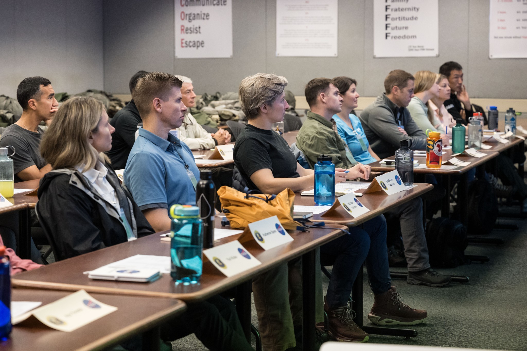 A classroom-style training session where people sit at tables with nameplates, notebooks, and water bottles, listening to an instructor, with motivational posters on the walls behind them.